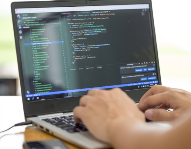 Close-up of a person typing code on a laptop displaying the vicidial auto dialer installation programming interface with multiple lines of code and dropdown suggestions. The image shows a bright and clear screen with a blurred background and a smartphone on the table next to the laptop.